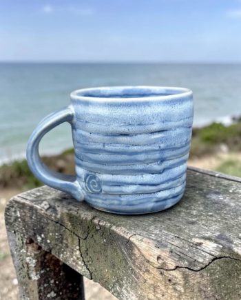 Lake Michigan ceramic mug with layered blue glaze and shoreline texture, shown overlooking the water