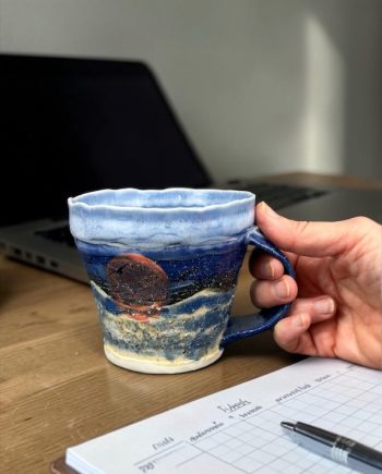 Handmade ceramic mug held in hand, showing sun motif, layered blue glaze, and aqua interior inspired by Lake Michigan