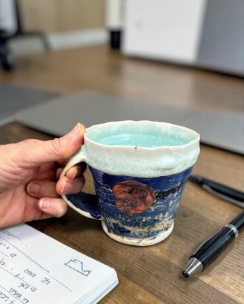 Ceramic mug held in hand, showing scale, sun detail, and layered glaze inspired by Lake Michigan shoreline
